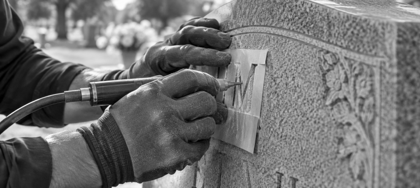 Gloved hands carving a letter through a stencil on a granite headstone